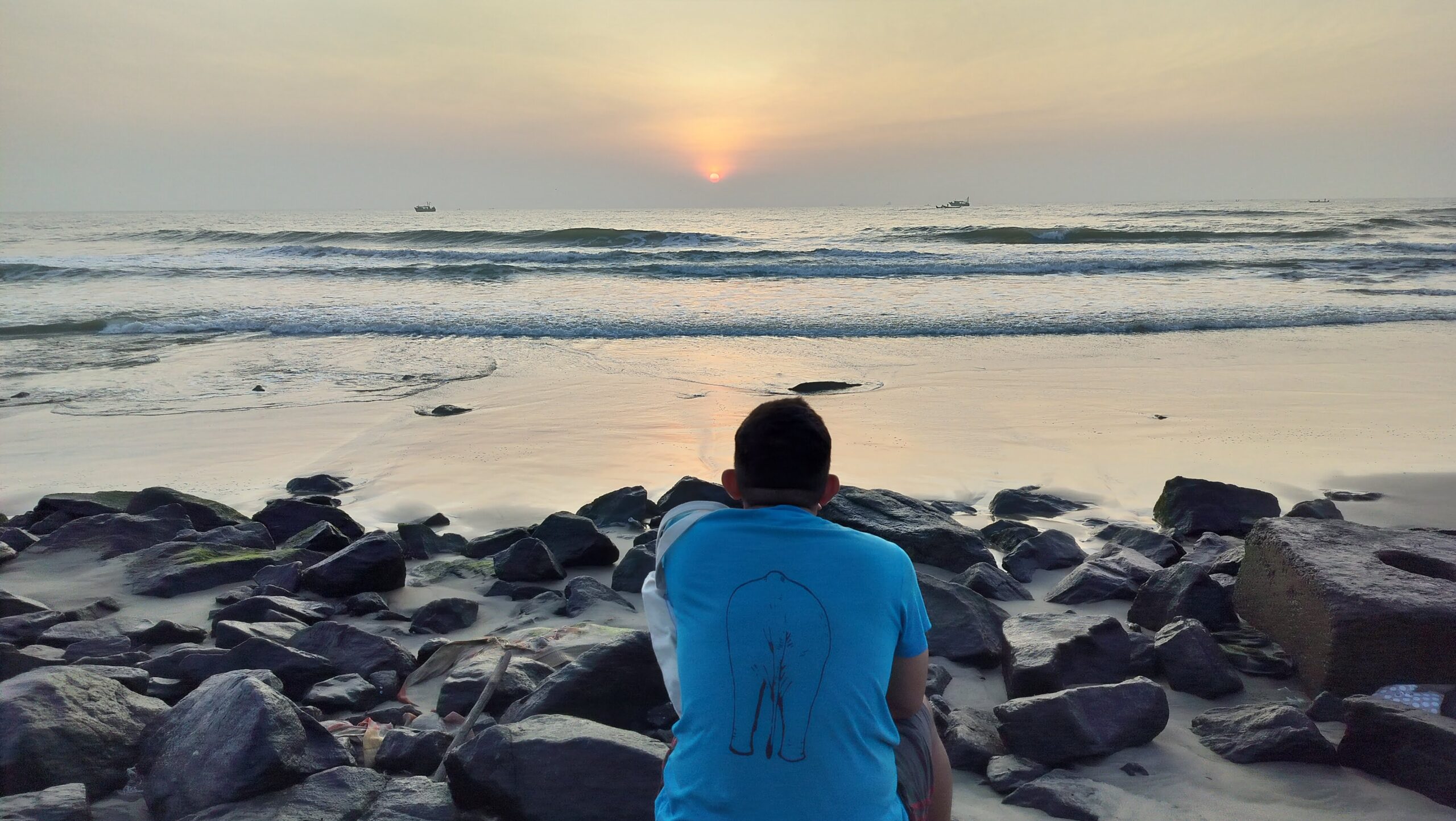 person in blue t-shirt looking at sunrise on a rocky beach